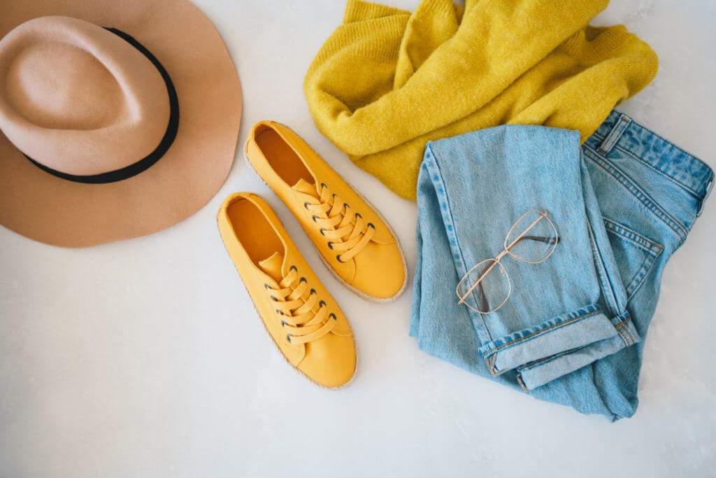 A series of clothes are laid out on a bed, including blue jeans, a yellow sweater, yellow shoes and a tan brimmed hat.