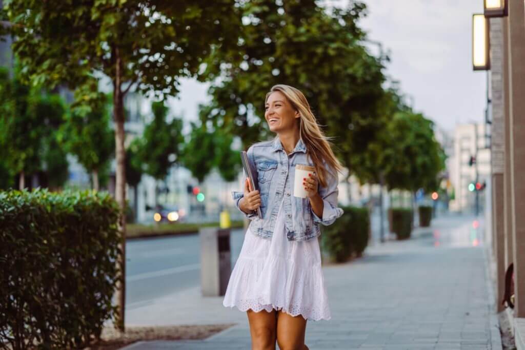 A woman walks down the street wearing a lavender sundress and a jean jacket.